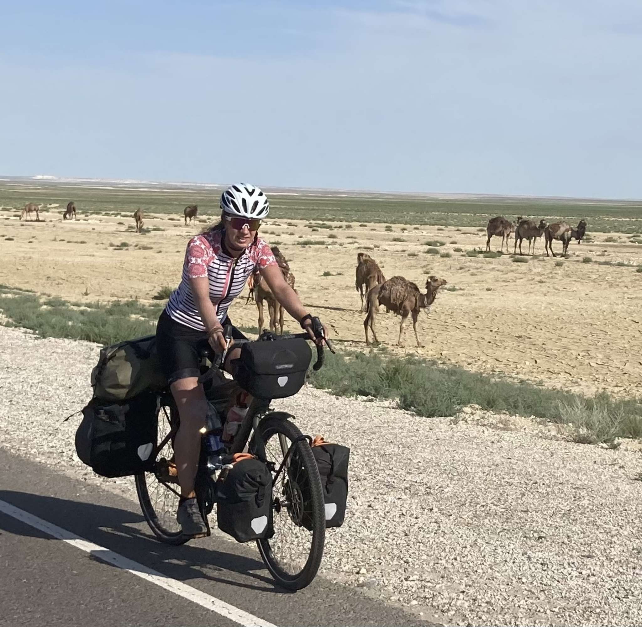 Marleen, chargée de bagages, roule sur une route à travers une steppe aride avec des chameaux en arrière-plan.